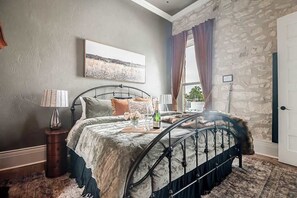 A bedroom with a stone wall, a bed with a metal frame, a nightstand, a lamp, and a framed picture on the wall. - The Bank on Main St - Downtown Escape Featuring a Deck w/Main St Views! (Fredericksburg)