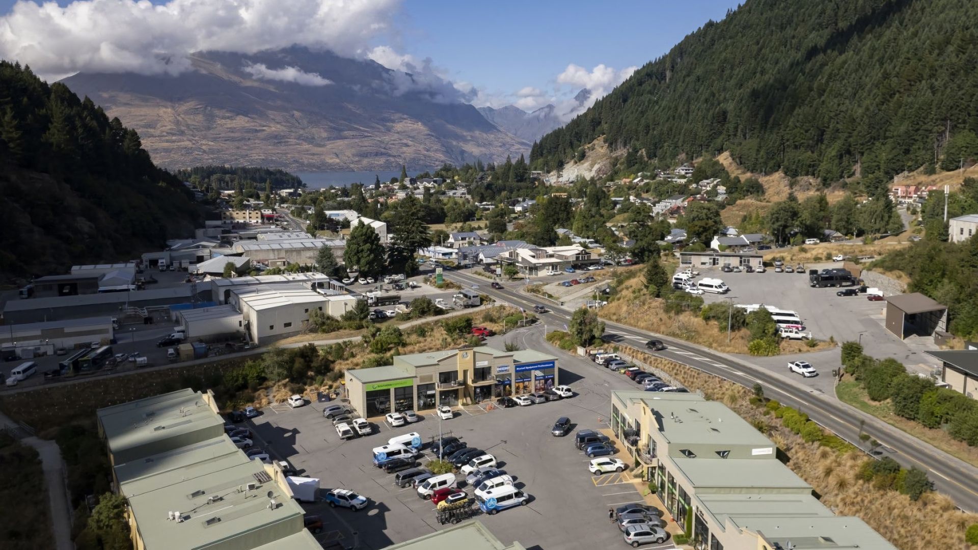 Une zone commerciale dotée d’un parking, de bâtiments et bordée par des montagnes.