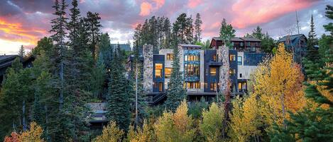 A multi-story lodge with a stone facade, surrounded by dense forest and autumn foliage.