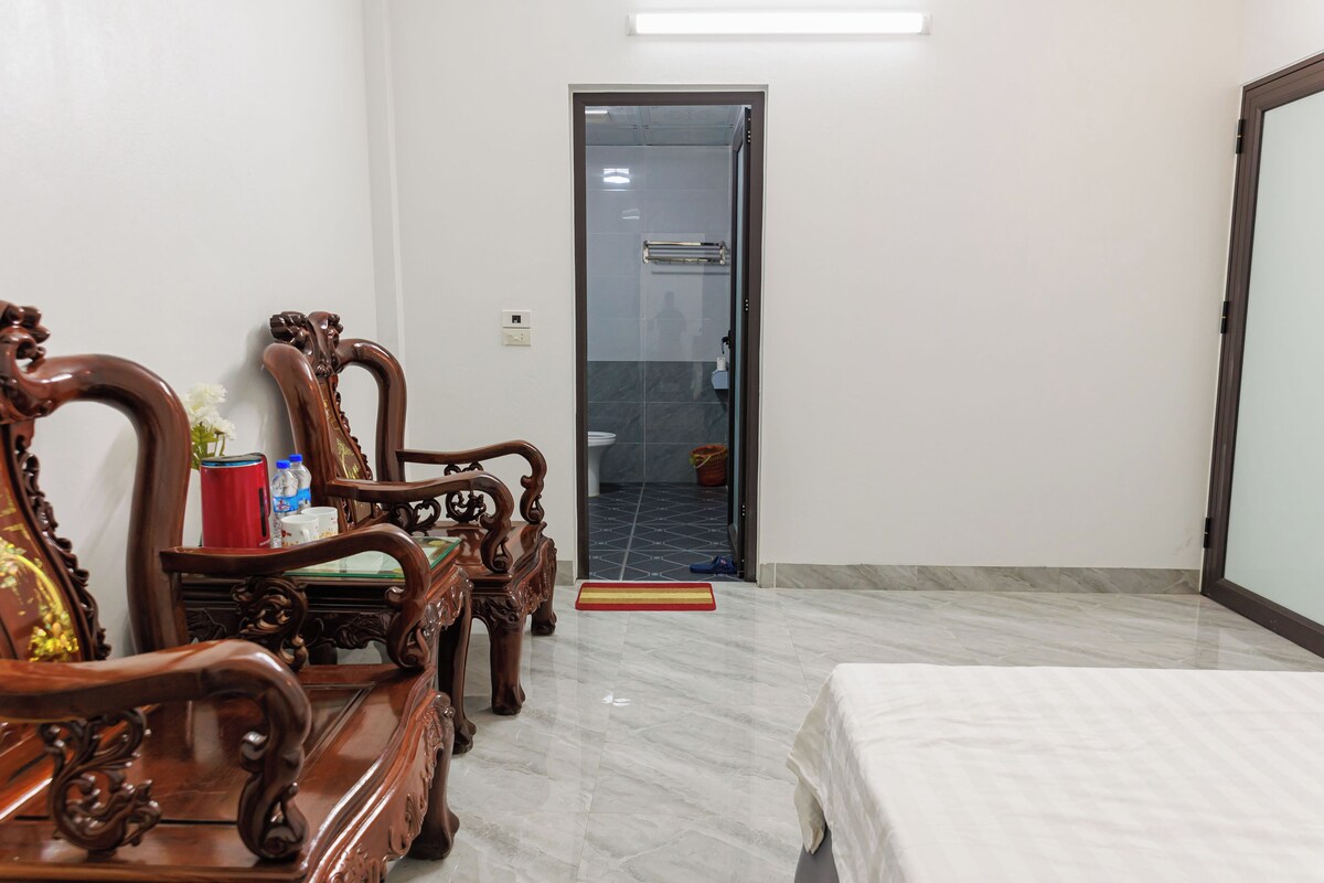 A room with a bed, wooden chairs, a small table with items, a glass door, and a tiled floor.