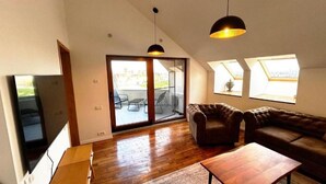 A spacious living area with a wooden floor, a brown leather sofa, a wooden coffee table, and a television. There are large windows with skylights and a sliding glass door leading to an outdoor patio. - The Admiral / Luxury Apartment in Constanta (Unknown)