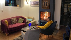Ein Wohnzimmer mit Kamin, einer Couch, einem Couchtisch mit einem Laptop und einer Person, die in einem Sessel sitzt.