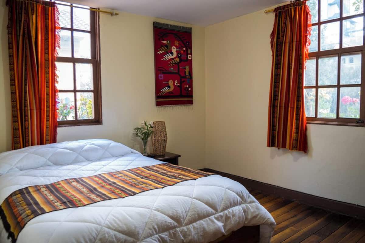 A bedroom with a large bed, two windows with curtains, a wall hanging, and a small table with a plant.