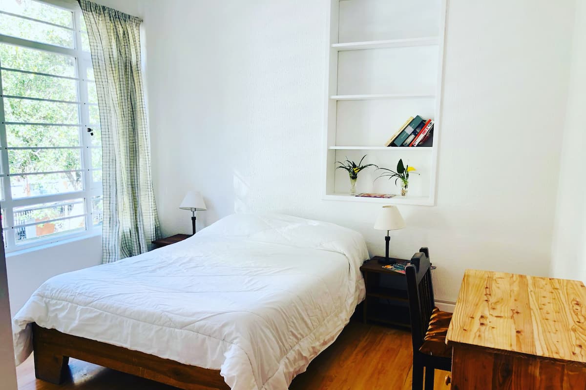 A neatly made bed with a white duvet, a bedside table with a lamp, a wooden nightstand, and a shelf with books and plants.