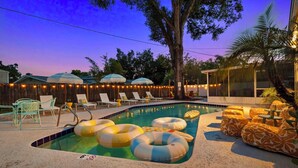 A swimming pool with lounge chairs and umbrellas, surrounded by string lights and trees. - 5BR~Huge Backyard~Minigolf~Heated Pool~Outdoor Projector~Gameroom~FIOS WiFi (Brandon)