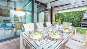 An outdoor dining area with glass tables, wicker chairs, and a view of a garden and house through large windows.