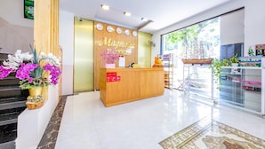A hotel reception area with a wooden reception desk, a digital clock, and a window with a view of greenery.