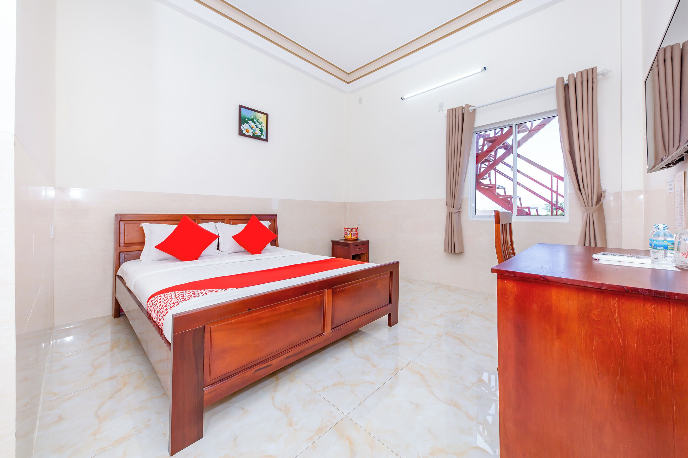 A hotel room with a wooden bed, a desk, a nightstand, a window with curtains, and a painting on the wall.