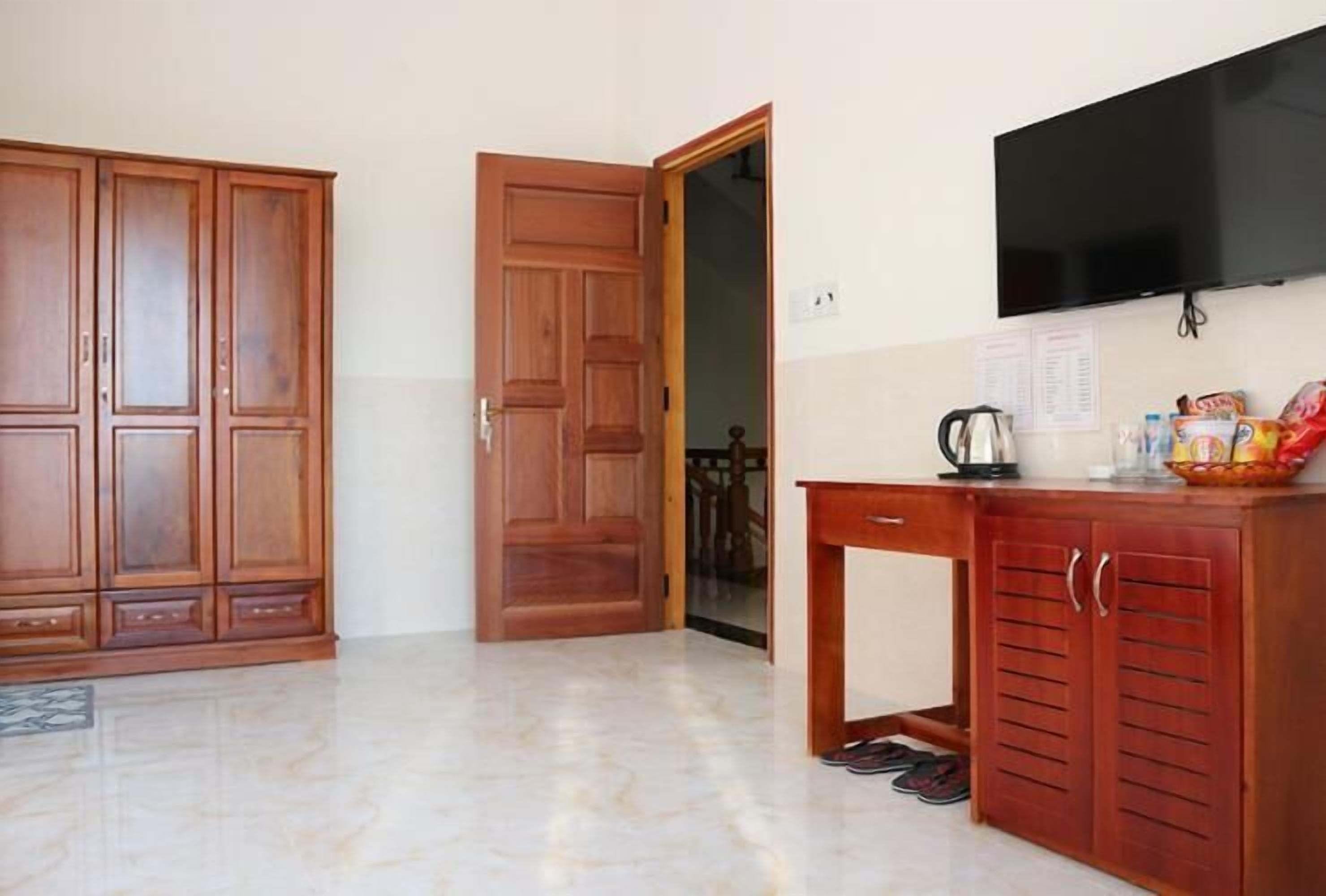 A room with a wooden wardrobe, a wooden desk with a television, and a tiled floor.