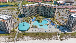 A large hotel with multiple floors, a central swimming pool, and a landscaped area with palm trees.