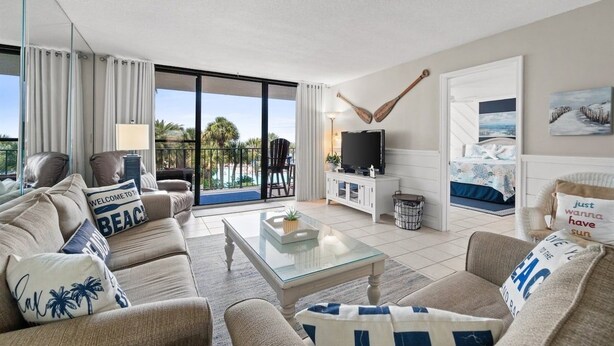 A living room with a sofa, coffee table, and a television. There is a sliding glass door leading to a balcony with a view of palm trees.