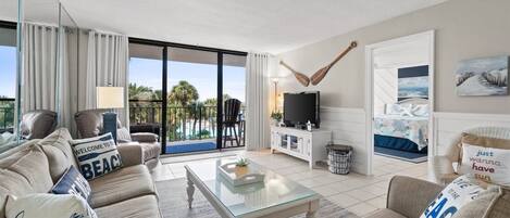 A living room with a sofa, coffee table, and a television. There is a sliding glass door leading to a balcony with a view of palm trees.