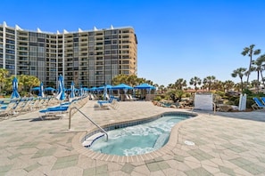 Área de piscina de hotel com espreguiçadeiras e guarda-sóis, uma banheira de hidromassagem e um edifício grande ao fundo.