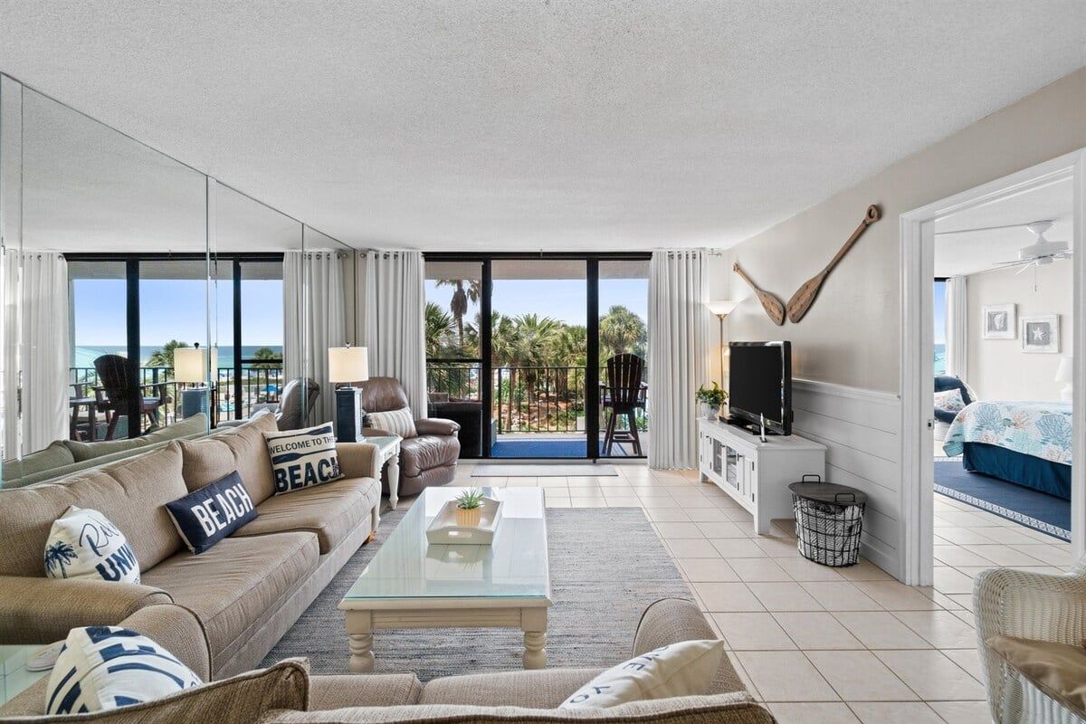 Una sala de estar moderna con un sofá, una mesa de centro y un televisor. Hay un balcón con vistas a palmeras y al océano.