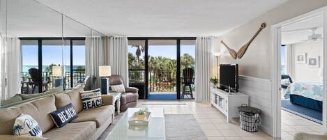 Una sala de estar moderna con un sofá, una mesa de centro y un televisor. Hay un balcón con vistas a palmeras y al océano.