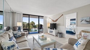 Ein Wohnzimmer mit Sofa, Couchtisch, Fernseher und Blick auf Palmen sowie einen Balkon.