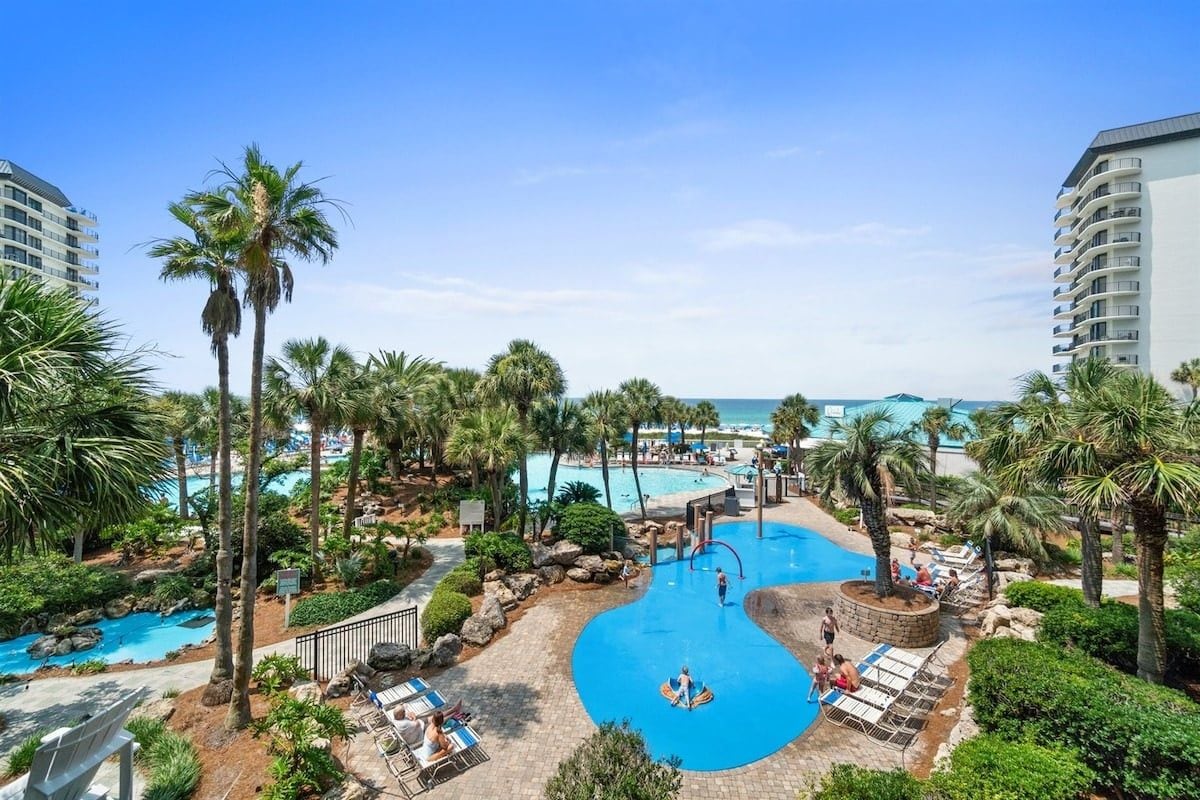 A resort with a central swimming pool, lounge areas, and palm trees.