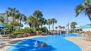 A swimming pool with a blue water feature, surrounded by palm trees and a building with a green roof.