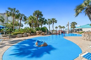 A swimming pool with a blue water feature, surrounded by palm trees and a building with a green roof.