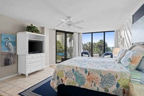 A bedroom with a bed, a TV, a ceiling fan, a balcony, and a view of palm trees.