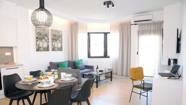 A modern living room with a sofa, dining table, and chairs. There is a TV, a laptop, and a window with curtains.