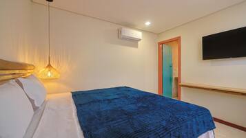 A hotel room with a bed, a hanging light fixture, an air conditioning unit, a television mounted on the wall, and a door with a teal frame.