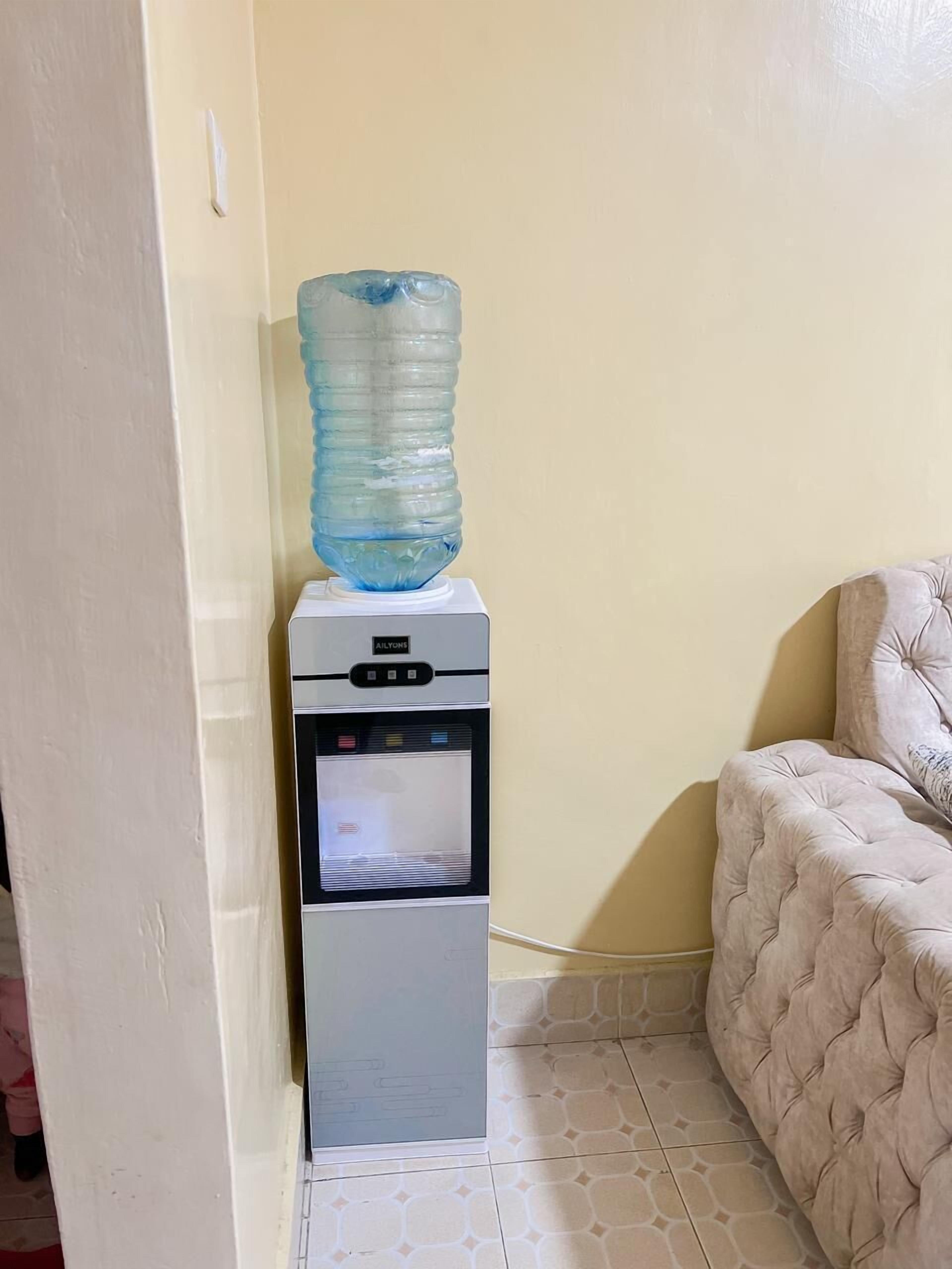 Dispensador de agua con una botella grande encima, ubicado en una esquina de habitación con pared color beige y un sofá capitonado.