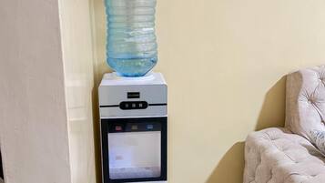 Dispensador de agua con una botella grande encima, ubicado en una esquina de habitación con pared color beige y un sofá capitonado.