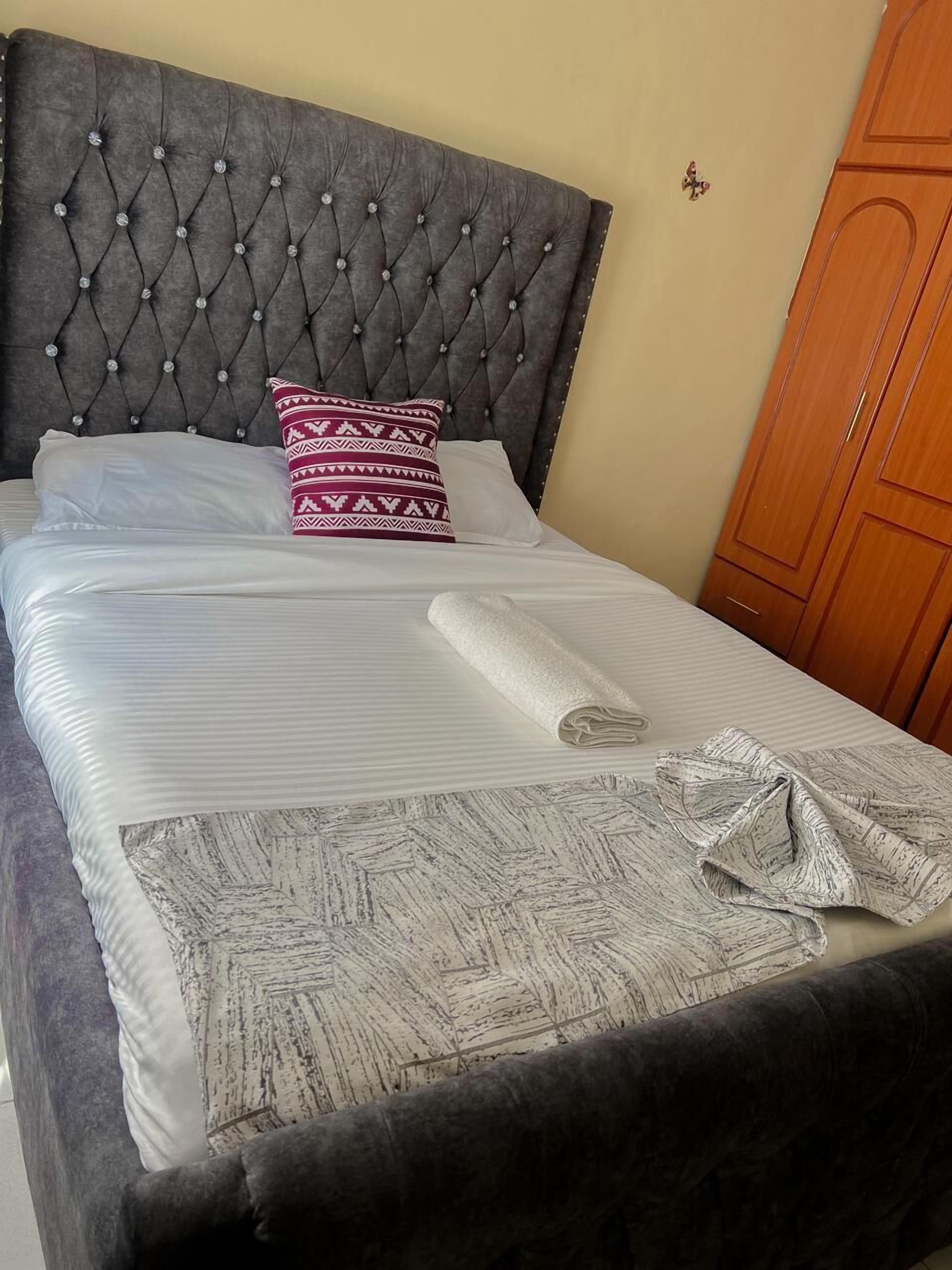 A neatly made bed with a tufted headboard, a patterned pillow, and a rolled towel on the bed.