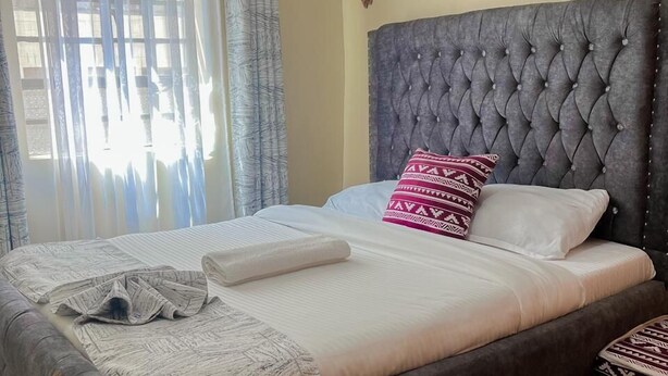 A neatly made bed with a tufted headboard, a patterned throw blanket, and a decorative pillow. - Lux Suites Cabre Apartments (Nakuru)