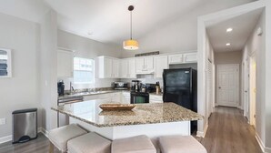 A modern kitchen with white cabinets, a granite countertop, and a black refrigerator. There are three bar stools with cushioned seats.