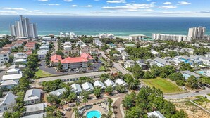 An aerial view of a coastal residential area with a large building featuring a swimming pool, surrounded by smaller houses and lush greenery.