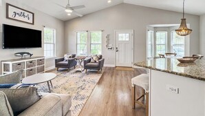 A spacious living room with a large TV, comfortable seating, and a kitchen area with a granite countertop.