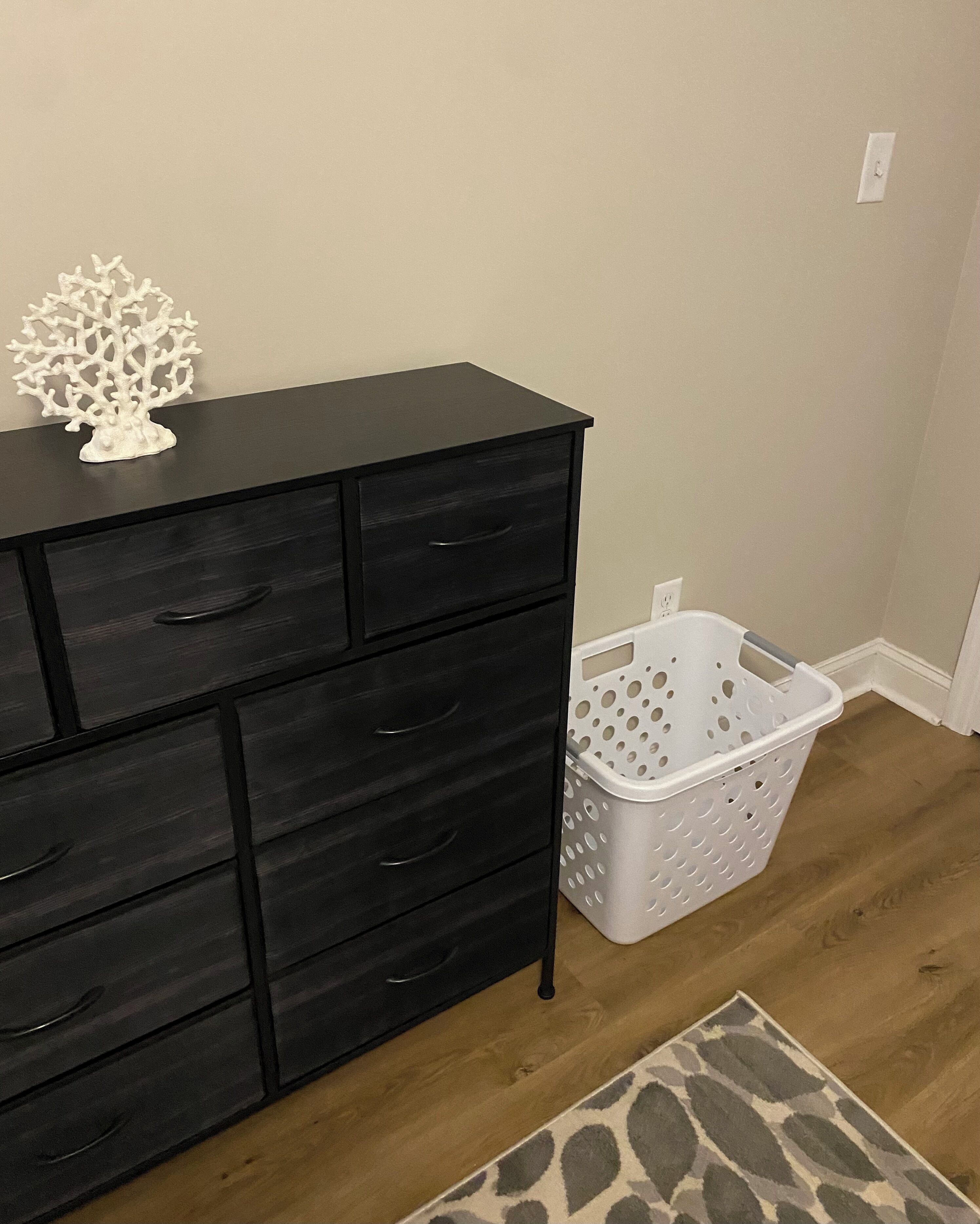 A black dresser with multiple drawers, a white decorative tree, a white laundry basket, and a patterned rug on a wooden floor.