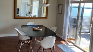 Een eetruimte met een donkere tafel, witte stoelen, een hangende lamp, een grote spiegel en uitzicht op de omgeving door een glazen deur.