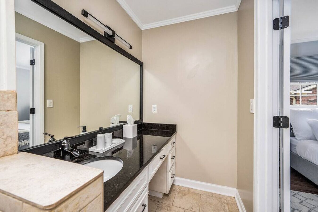 A modern bathroom with a large mirror, a black countertop, a sink, and a door leading to a bedroom.