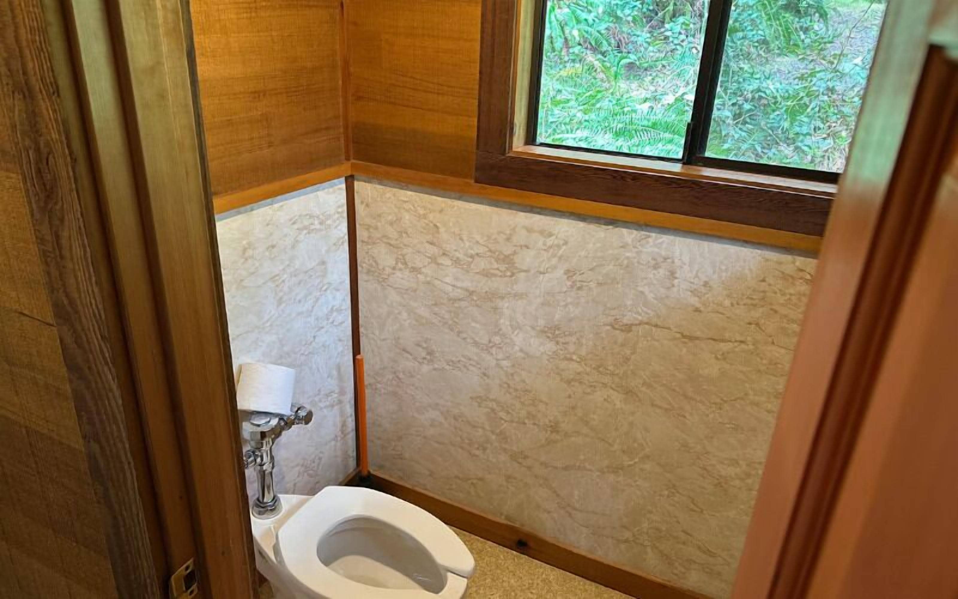 A small bathroom with a toilet, marble walls, and a window with a view of greenery.