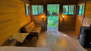 A cozy cabin interior with a sofa, a bed, a small table, and a refrigerator. There are two windows with lights on, and a door leading to a forested area. - Smith Creek Village  (Sublimity)