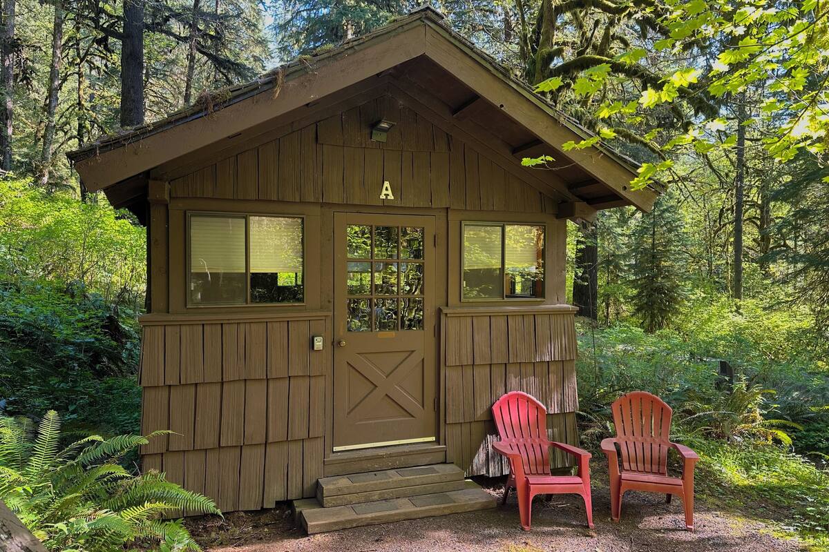 A small wooden cabin with a door marked 'A', two red Adirondack chairs, and a gravel path leading to the entrance.