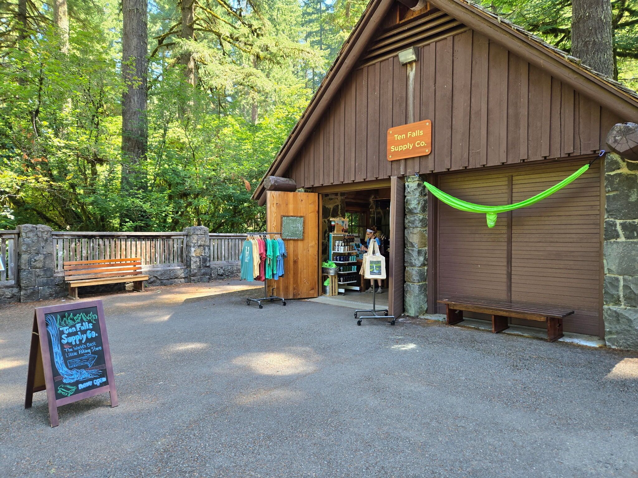 A wooden building labeled "Ten Falls Supply Co." with a signboard, a bench, and a rack of colorful shirts outside.