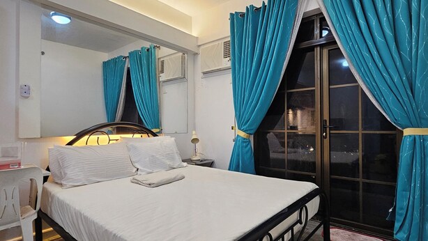 A bedroom with a bed, a bedside table, a mirror, and a window with curtains. - Cozy 1-bedroom condo with AC and WiFi in central Davao City (Davao City)