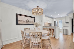 A dining area with a round table, wicker chairs, a chandelier, and a framed cityscape picture on the wall. - Award-Winning Resort Amenities | Racquet Club Villa 15 (Isle Of Palms)