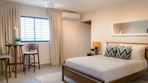 A bedroom with a wooden bed, a desk with a chair, a window with curtains, an air conditioning unit, and a bedside table with a lamp and decorative items.