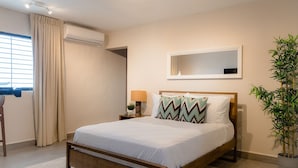 A bedroom with a wooden bed, a bedside table with a lamp, a potted plant, and a window with curtains.