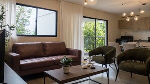 A modern living room with a brown leather sofa, a wooden coffee table, and a green armchair. There is a large window with curtains, a kitchen area in the background, and a ceiling light fixture.