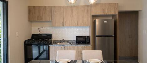 A modern kitchen with wooden cabinets, a stainless steel refrigerator, a microwave, and a gas stove. There is a dining table set with plates, glasses, and cutlery.