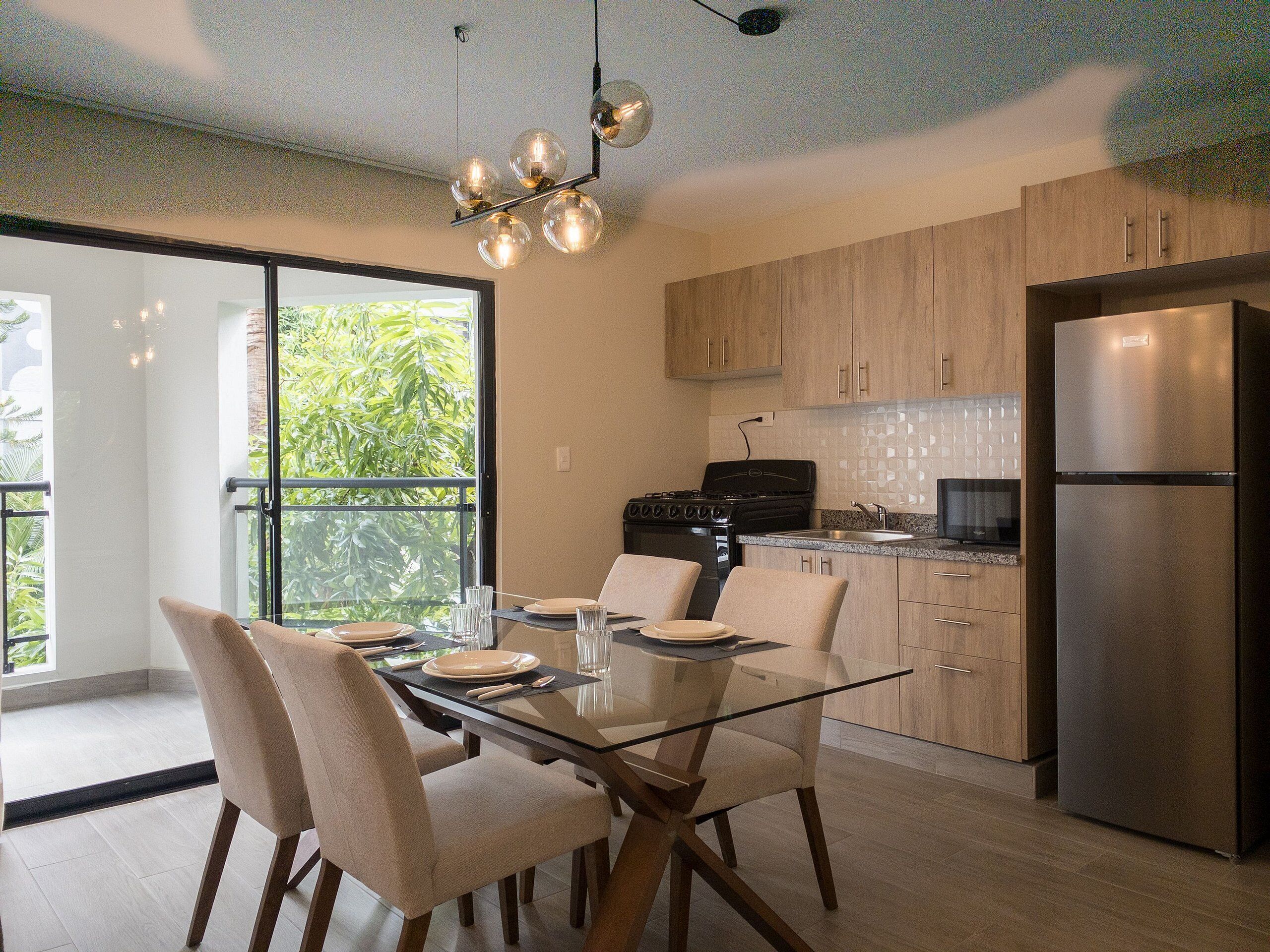 A modern kitchen with a glass dining table, wooden cabinets, and a stainless steel refrigerator.