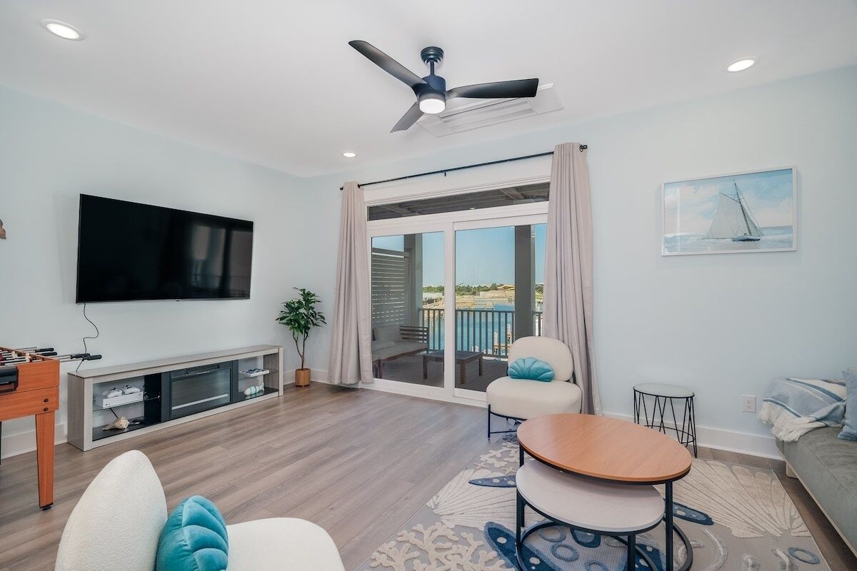 A modern living room with a flat-screen TV, a ceiling fan, a sofa, a coffee table, and a balcony with curtains.