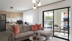 A modern living room with a grey sofa, two pink cushions, and a coffee table with decorative vases. A kitchen with wooden cabinets and a dining area with a table and chairs are visible in the background. - Modern 2BR ·Gazcue · 5min to Monuments · YellowKey (Santo Domingo)
