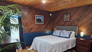 Une chambre avec un plafond en bois, un lit recouvert de linge de lit blanc, une table de chevet avec une lampe, une grande plante et un tableau encadré au mur.